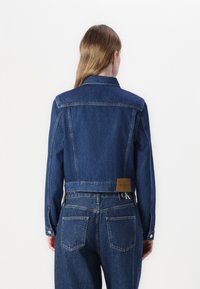 Giacca cropped in denim blu scuro con colletto, cuciture tradizionali e un'etichetta marrone sulla vita sul retro. Presenta chiusure con bottoni classici.