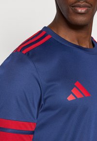 Blaues Sportshirt mit roten Akzenten an Schultern und Ärmeln. Das Material ist glatt und verfügt über einen Rundhalsausschnitt. Vorderseite mit Logo versehen.