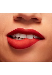 Rossetto opaco rosso applicato su labbra piene, che mostra una superficie liscia e un contorno definito. Denti bianchi visibili tra labbra leggermente aperte.