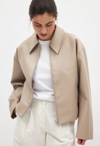 Beige vadderad jacka med spetsig krage och framdragkedja. Har sänkta axlar och vida ärmar, bärs över en vit t-shirt.