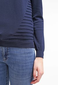 Marineblauer Strickpullover mit gerippten Bündchen und seitlichem Design, das horizontale Aussparungslinien aufweist. Kombiniert mit blauen Jeans.