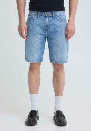 Mężczyzna w jasnoniebieskich krótkich spodenkach dżinsowych, białych skarpetkach i czarnych mokasynach, stojący na tle jasnego, jednolitego tła.