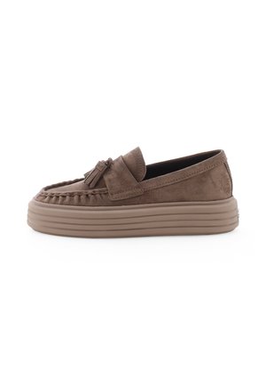 Bruine suède loafer met dikke geribbelde zool en kwastdetail bovenop, van opzij getoond op een witte achtergrond.
