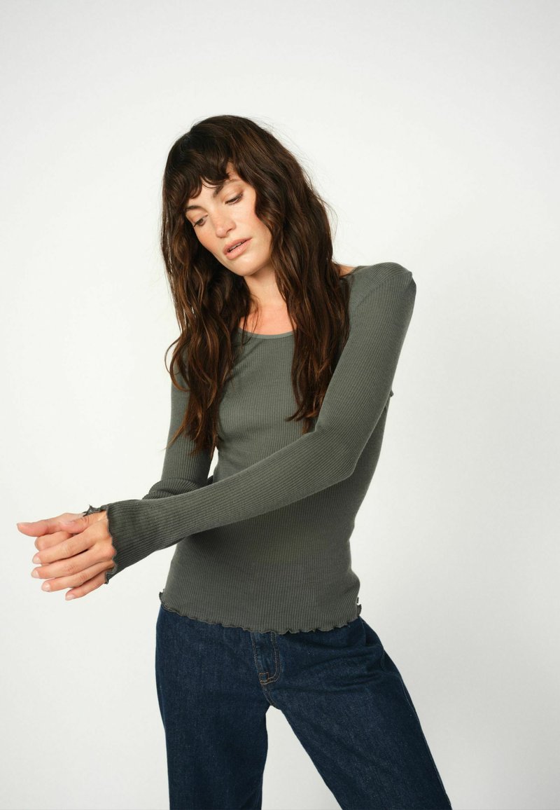 Donkergroene top met lange mouwen en ribbelstof, voorzien van een ronde halslijn en rafelige manchetten, gecombineerd met donkerblauwe jeans. Stevig ontwerp met gestructureerde stof.