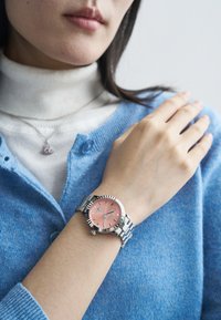 Ασημένιο ρολόι από ατσάλι με ροζ καντράν, διαθέτει ασημένιους δείκτες ώρας και ραβδωτό πλαίσιο, φορεμένο σε καρπό με μπλε πουλόβερ.