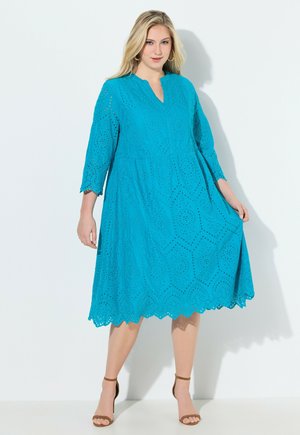 Kvinde iført turkisblonde midi-kjole med bølget kant og 3/4-ærmer, kombineret med brune ankelstrop-hæle, stående mod hvid baggrund.