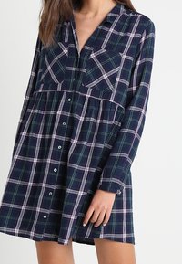 Vestido de cuadros de manga larga en navy, verde y blanco. Presenta un cierre frontal de botones, un cuello y dos bolsillos en el pecho. Forma tipo A con cintura fruncida.