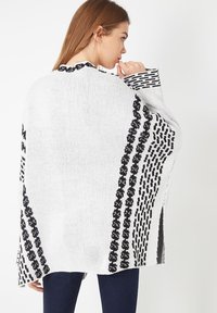 Mujer de perfil vestida con un suéter de punto blanco suelto con patrones geométricos negros y pantalones oscuros, contra un fondo blanco liso.