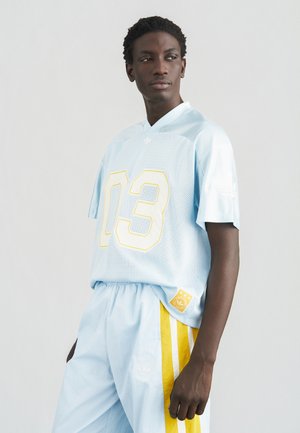 Camiseta de malla azul claro con el número "03" en amarillo llamativo y detalle de raya amarilla en los pantalones cortos azul claro, complementada con un parche con el logo.