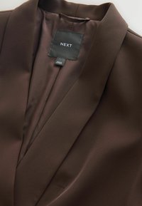 Barna zakó sima textúrával, állógallérral, belsejében fekete márka címkével, amelyen az "NEXT" és a "Made in Vietnam" felirat található.