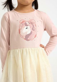 Helles rosa Langarmkleid mit einem Einhornmotiv und Pailletten am Oberteil; flauschiger cremefarbener Rocküberzug mit goldenen Sprenkeln.