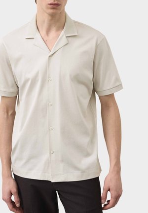 Man draagt een beige overhemd met korte mouwen, een gebreide kraag en geribde manchetten, gecombineerd met een zwarte broek, tegen een effen achtergrond.