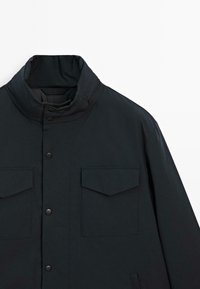 Giacca blu scuro con colletto alto, due tasche sul petto con pattina e bottoni a pressione. Realizzata in tessuto morbido con un design leggermente strutturato.