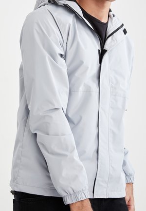 Hombre que lleva una chaqueta cortavientos con capucha de color gris claro sobre una camiseta negra, de pie frente a un fondo blanco.