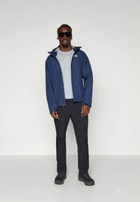 Navy waterproof jacket with a hood, paired with a light gray shirt and dark pants; worn with sunglasses and black hiking shoes.