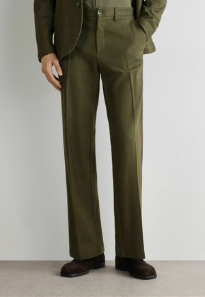 Mann trägt olivgrüne Stoffhose, dazu passenden Blazer und dunkelbraune Wildlederschuhe, steht vor einer schlichten hellen Wand.