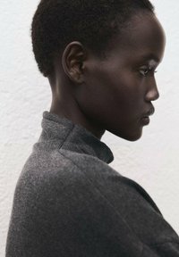 Siv pulover z teksturo in visokim ovratnikom, viden od strani. Ima kratko pristrižene lase in gladko kožo na svetli podlagi.