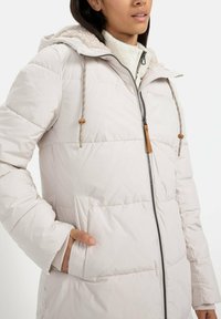 Helle Beige Daunenjacke mit Reißverschluss vorne, einem mit Fell gefütterten Kapuze, Kordelzug und Seitentaschen. Verfügt über eine gesteppte Textur und einen kontrastierenden Reißverschluss.