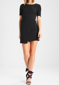 Svart klänning med prickar, korta ärmar, figursydd design och omslagsfront. I kombination med lila sandaler med remmar och snörningar.