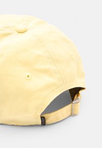 Gul bomulds cap med en tekstureret overflade, seks-panel design, justerbar rem og et lille mærke på siden. Har topknap og øjenhuller.