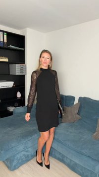 Czarna sukienka z cekinowymi długimi rękawami, dopasowana sylwetka. Modelka nosi czarne szpilki, stoi na teksturowanej niebieskiej sofie. Neutralne tło.