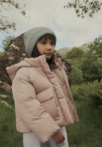 Gefütterte, kurze Jacke in hellrosa mit Kapuze, ausgestattet mit zwei Fronttaschen und einer glatten Textur. Begleitet von einer grauen gestrickten Mütze.