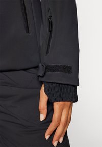 Zwarte softshell jas met een mouw van zacht materiaal, een geperforeerde elastische manchette, klittenbandsluiting en twee ritszakken op de arm, met een gladde afwerking.