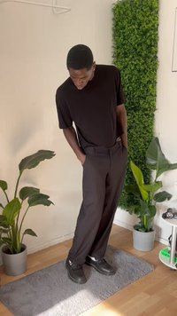 Black short-sleeve t-shirt, relaxed-fit black trousers, and black shoes. Room features green plants and a gray rug on wooden floor.
