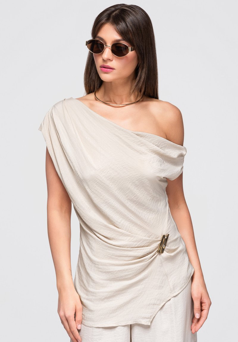 Femme aux cheveux bruns raides porte une robe beige épaules dénudées avec une broche dorée, des lunettes de soleil foncées et un fin collier en or sur un fond uni.