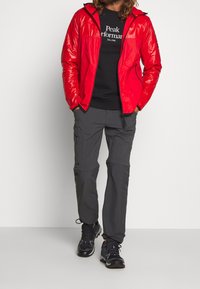 Rød, skinnende jakke med lynlås og hætte over en sort t-shirt; grå cargobukser med lommer; sorte vandresko.