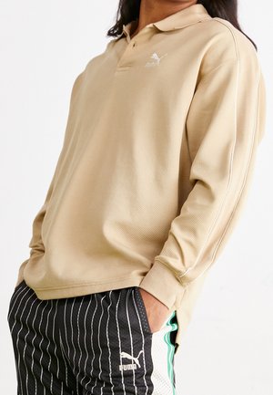 Polo a maniche lunghe beige con motivo testurizzato, caratterizzato da una patta con tre bottoni e logo Puma bianco sul petto sinistro, abbinato a pantaloni neri a righe.