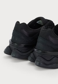 Paire de baskets noires New Balance montrant des talons rembourrés, des semelles épaisses rainurées et un logo à l'arrière sur fond blanc.