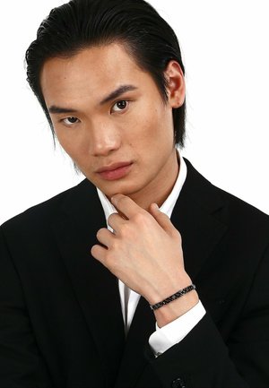 Zwarte armband met een gestructureerd design, gedragen om de pols van een persoon in een zwart pak en een wit overhemd, staande tegen een witte achtergrond.