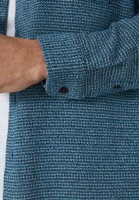 Personens håndled og hånd justerer ærmet på en tekstureret blå knapjakke over en hvid skjorte.