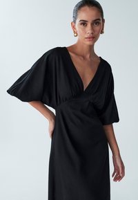 Black dress with a deep V-neck, short voluminous sleeves, and a fitted waist. Made of smooth fabric, it falls to a mid-calf length.