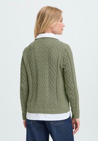 Gebreide trui in olijfgroen met kabelpatroon en geribde manchetten. Gelaagd over een witte blouse met kraag, met een ontspannen pasvorm.