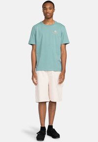 Camiseta de algodón azul claro con escote redondo y logo naranja. Combinada con pantalones cortos cargo beige. Zapatos negros con una superficie texturizada.
