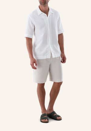 Mann trägt weißes kurzärmeliges Hemd mit Knöpfen, hellbeige Shorts und dunkelgrüne Sandalen, steht vor einfarbigem Hintergrund.