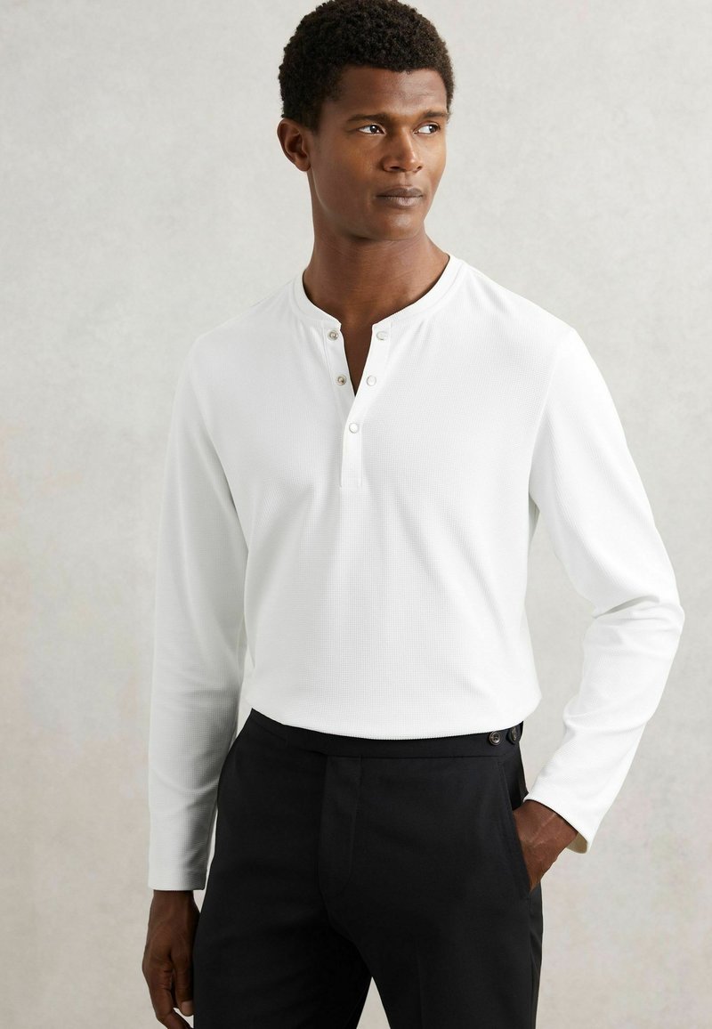 Weißes Langarm-Henley-Shirt aus strukturiertem Stoff, mit einem Drei-Knopf-Verschluss und Figur betonter Silhouette, kombiniert mit schwarzen Hosen.