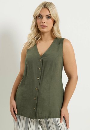 Donna bionda che indossa una tunica senza maniche verde oliva con bottoni e scollo a V, e pantaloni bianchi a righe, in piedi davanti a uno sfondo semplice.