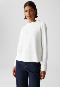 Vrouw met kort donker haar, gekleed in een textuurrijk wit long-sleeve shirt en donkerblauwe jeans, op een effen lichte achtergrond.