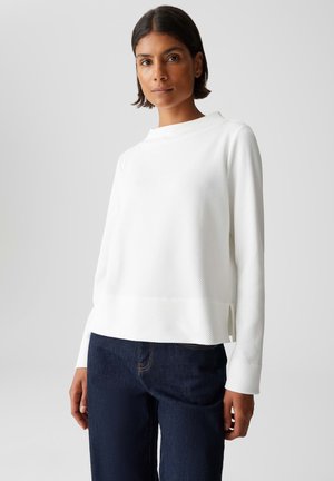 Vrouw met kort donker haar, gekleed in een textuurrijk wit long-sleeve shirt en donkerblauwe jeans, op een effen lichte achtergrond.