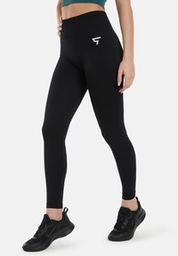 Črne legice visoke pasu z gladko teksturo, ki imajo majhen bel logo na eni stegnu in prilegajoč dizajn. Črne športne čevlje.