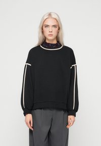 Μαύρη μπλούζα με υπερμεγέθη μανίκια, αντίθετη μπεζ διακοσμητική λεπτομέρεια γύρω από τον λαιμό και τους ώμους, μαλακό ύφασμα, σε συνδυασμό με γκρίζους παντελόνες με φαρδιά γραμμή.