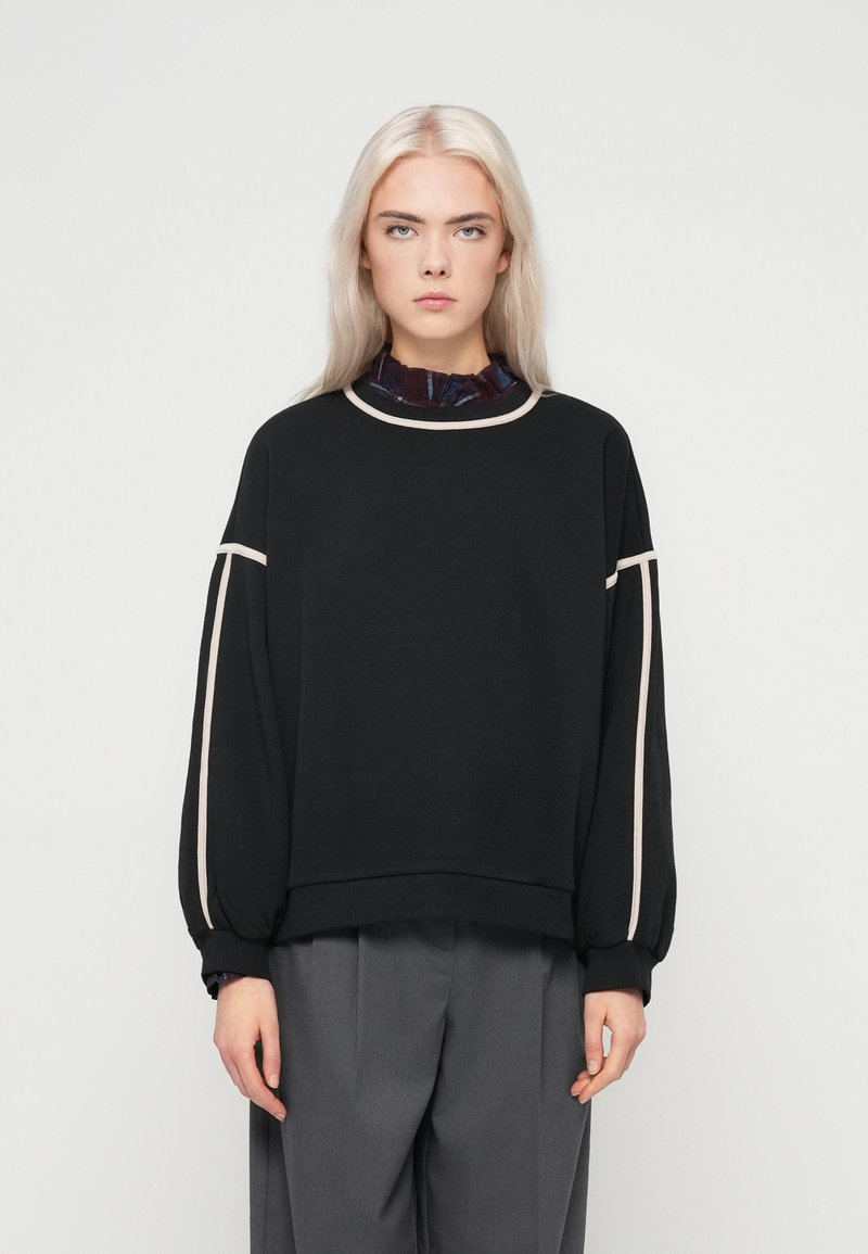 Μαύρη μπλούζα με υπερμεγέθη μανίκια, αντίθετη μπεζ διακοσμητική λεπτομέρεια γύρω από τον λαιμό και τους ώμους, μαλακό ύφασμα, σε συνδυασμό με γκρίζους παντελόνες με φαρδιά γραμμή.