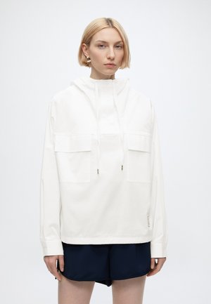 Femme blonde portant un sweat à capuche blanc avec des poches avant et un short bleu marine, debout devant un fond clair uni.