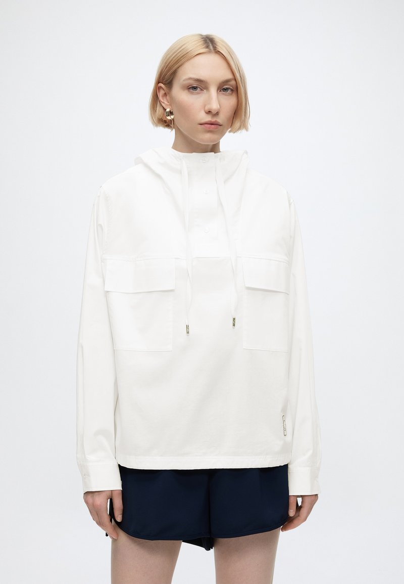 Femme blonde portant un sweat à capuche blanc avec des poches avant et un short bleu marine, debout devant un fond clair uni.