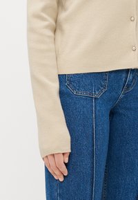 Beige cardigan med en myk tekstur, med en tettsittende design og perleknappdetaljer. Sammen med høy midje blå denimjeans.