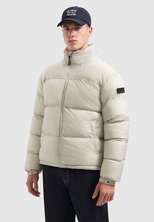 Pufferjack in lichtgrijs, met een hoge kraag, ritssluiting, zijzakken en een zwart logo-embleem op de mouw. G gedragen met donkere jeans.