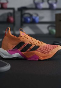 Chaussure de sport orange et rose avec des bandes noires sur le sol du gymnase, kettlebells flous en arrière-plan.